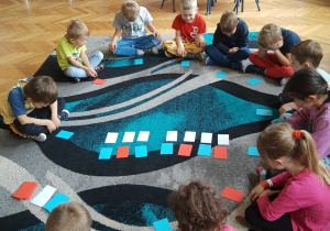 Dzieci przedszkole siedzą na dywanie w klasie w siadzie skrzyżnym. Uczestniczą w zajęciach w zakresie analizy i syntezy wyrazów z wykorzystaniem nakrywek w trzech kolorach; białym, czerwonym i niebieskich. Na zdjęciu widoczne skupienie i chęć współpracy podczas wykonywania zadania.